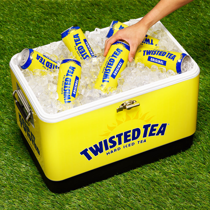 A bright yellow Twisted Tea hard iced tea cooler filled with ice and Twisted Tea Original cans, positioned outdoors on green grass, with a hand reaching into the cooler to grab a can.