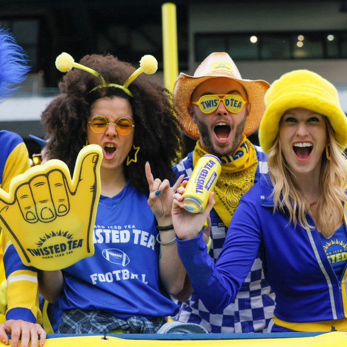 Three excited fans wearing bright yellow and blue Twisted Tea sports gear, including a foam finger, a can of Twisted Tea, a yellow bucket hat, and a checkered scarf, cheering at a football game.