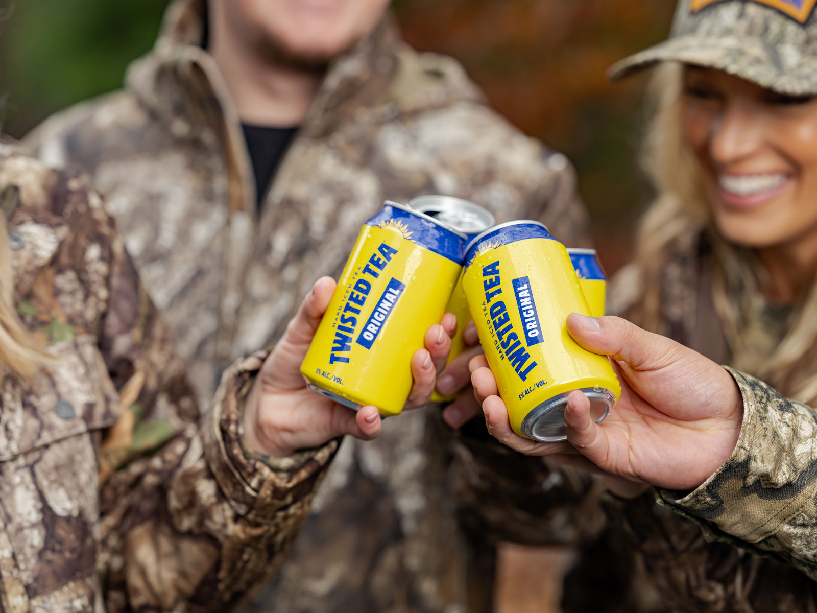 Two people in camouflage clothing clink yellow and blue Twisted Tea Canopy cans in a outdoor setting.
